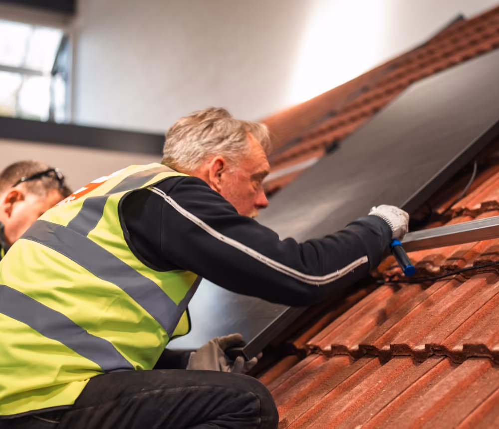 A worker in a high-visibility vest kneels on a tiled roof, fitting a solar panel.