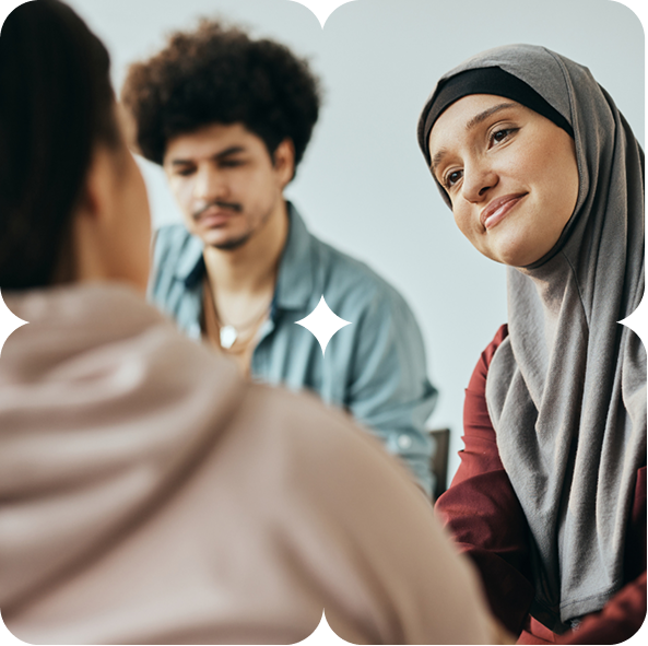 A woman wearing a hijab attentively listens to a person whose back is to the camera, with a man in the background.