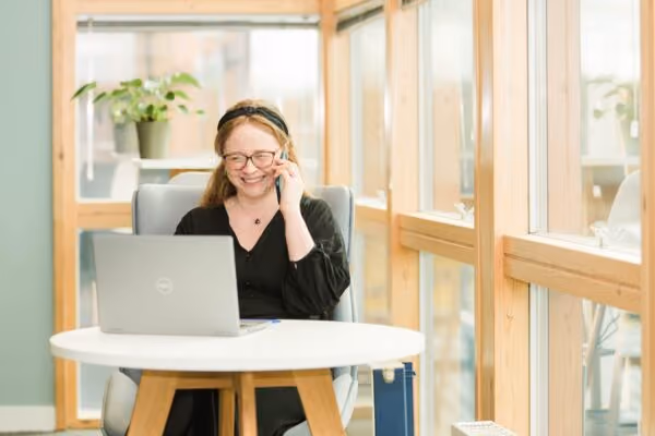 Smiling woman with glasses on phone, working at laptop in bright office space with windows.