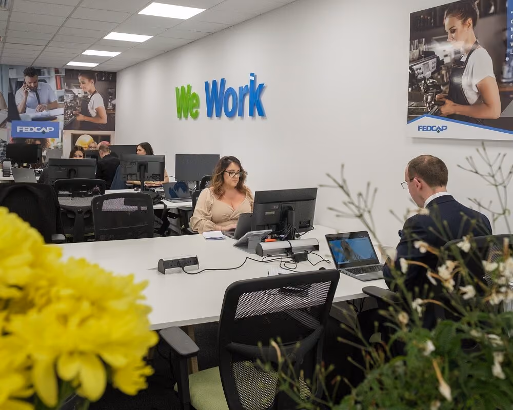 Office with people working at desks; wall has 'We Work' sign and Fedcap posters; flowers in foreground.