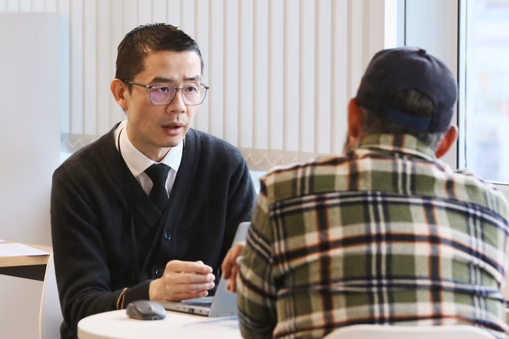 Man in glasses and tie talking to another man wearing a cap and plaid shirt at a round table.