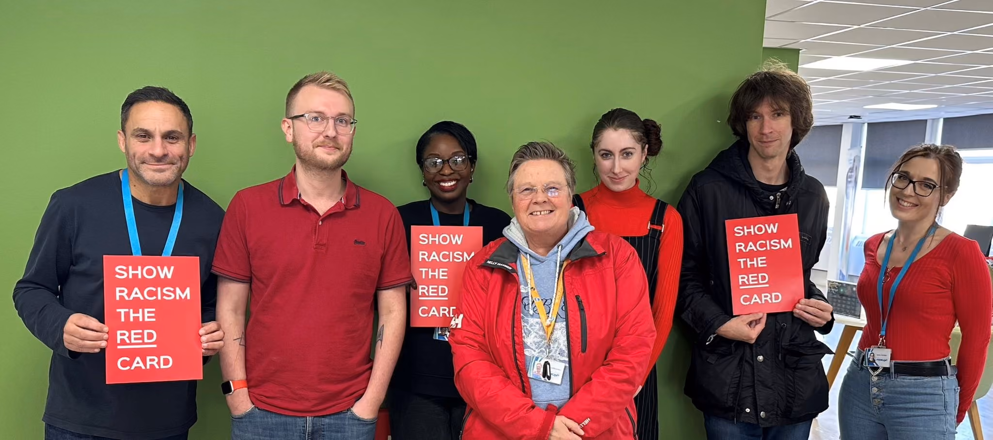 Seven colleagues pose together holding red “Show Racism the Red Card” signs in front of a green wall.