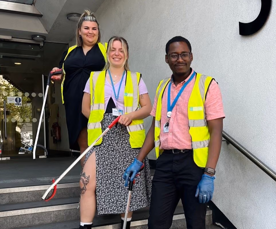 Three Fedcap volunteers wearing yellow hi-vis vests pose together holding litter pickers outside a building.