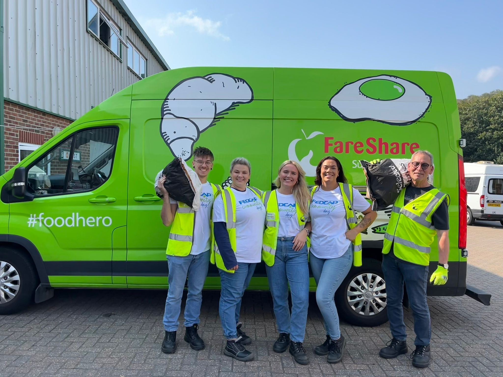 Six Fedcap volunteers wearing Fedcap t-shirts and hi-vis vests hold donation bags in front of a green FareShare food delivery van.
