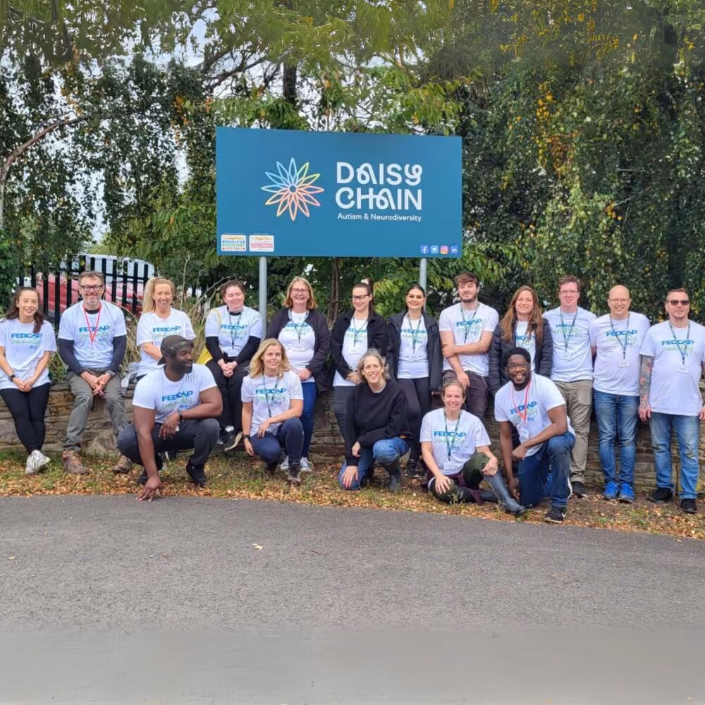 A large group of Fedcap staff in matching t-shirts pose together outdoors next to a Daisy Chain sign.