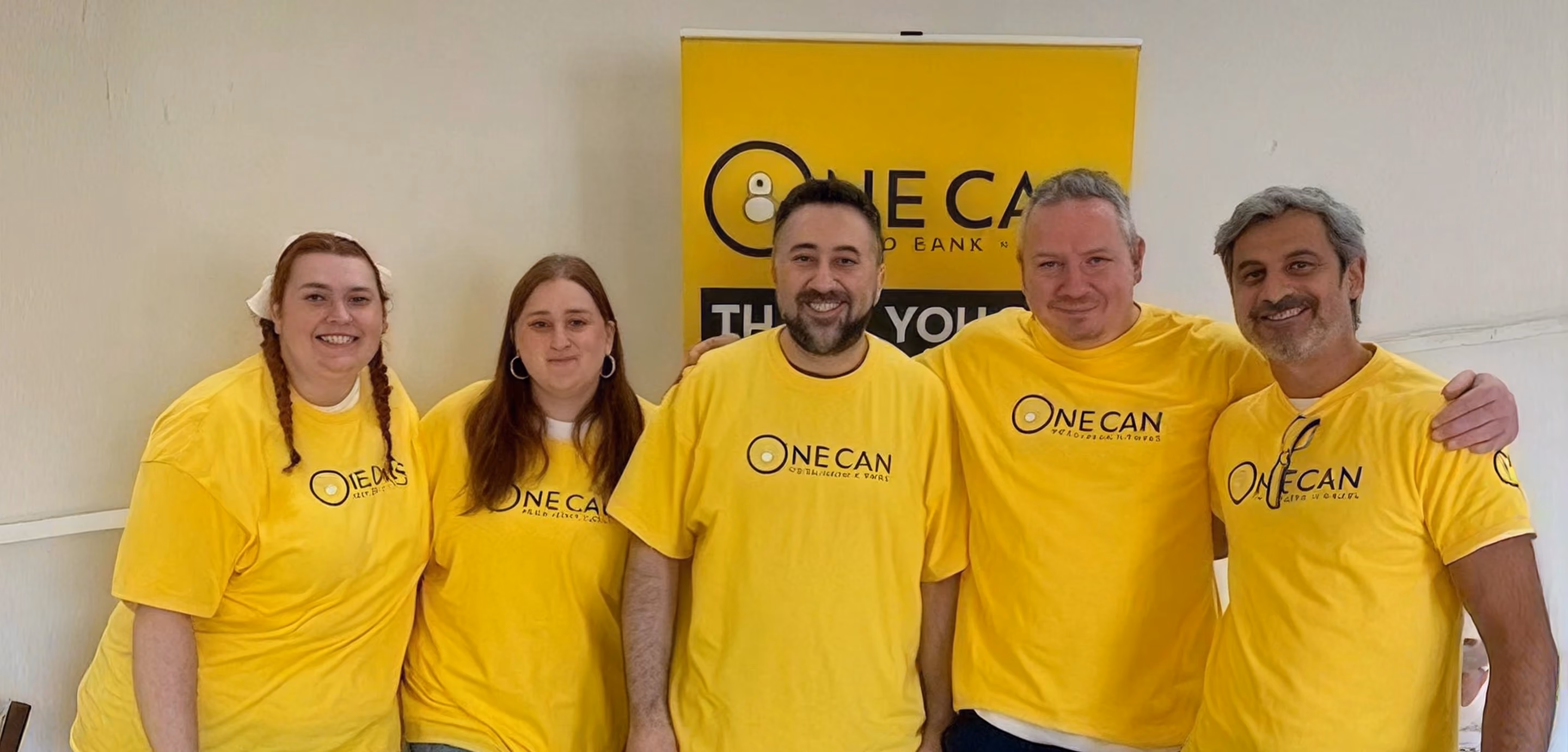Five volunteers in matching yellow One Can Food Bank t-shirts pose together indoors.