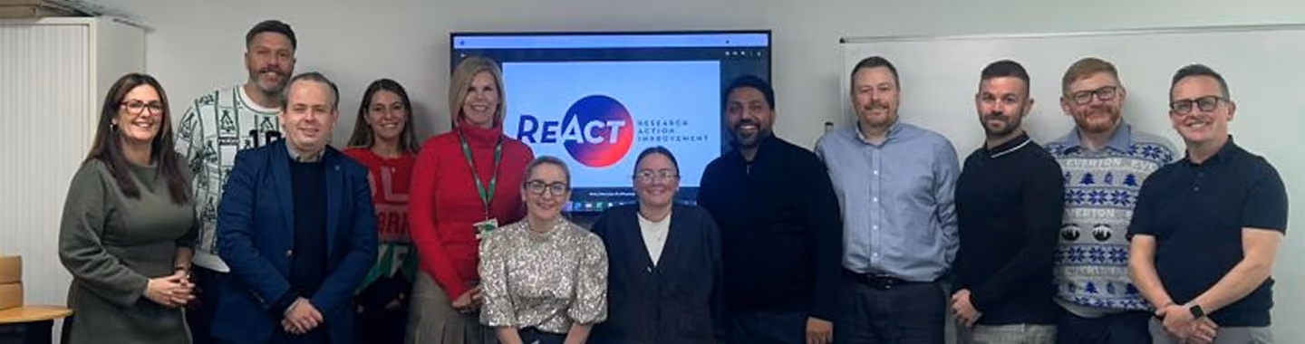 A group of Fedcap colleagues pose together in an office in front of a screen displaying the REAct (Research Action Improvement) logo.