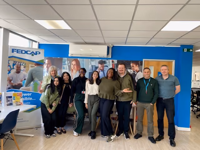 A group of ten Fedcap colleagues pose together for a photo in an office in front of a wall mural.