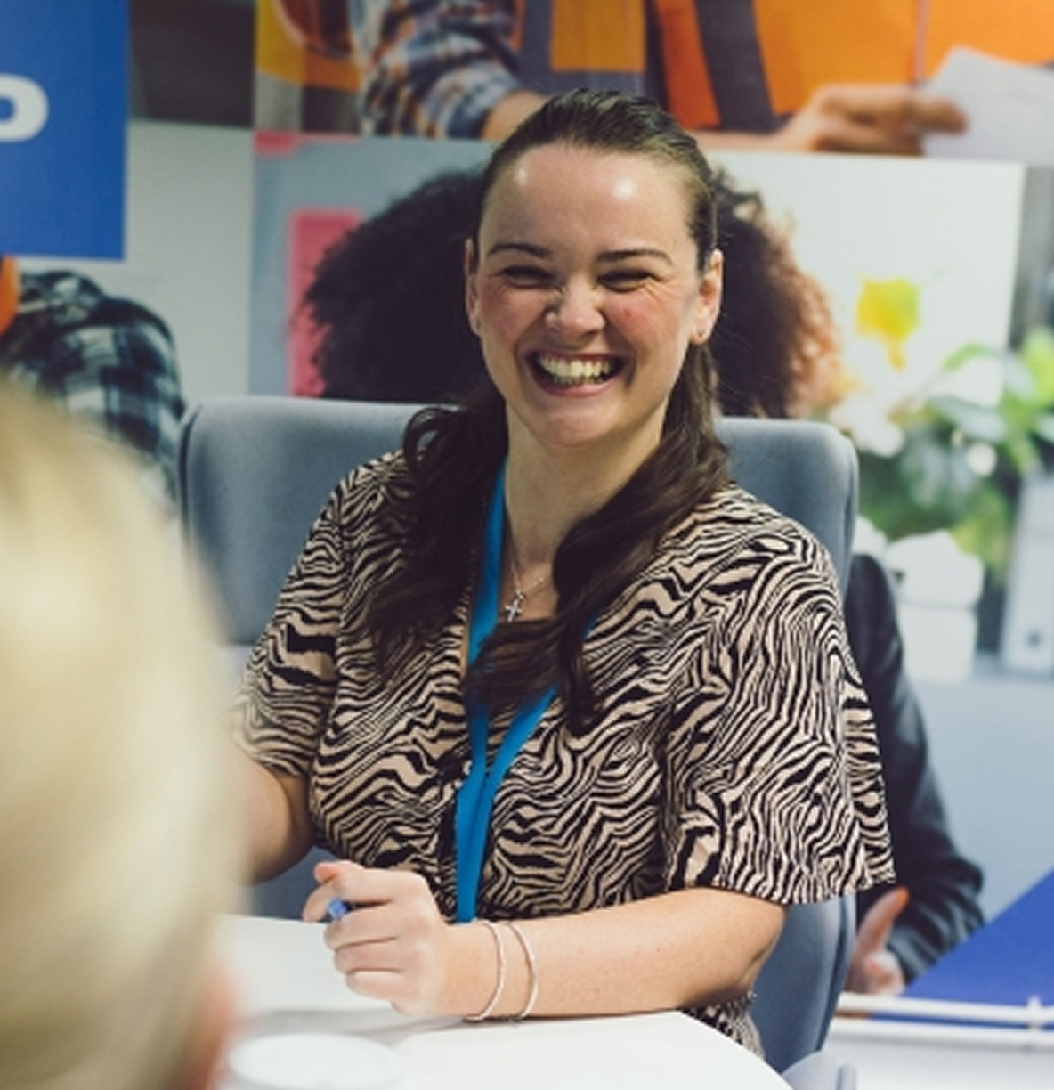 A smiling woman in a zebra-print top and blue lanyard sits at a table, holding a pen.