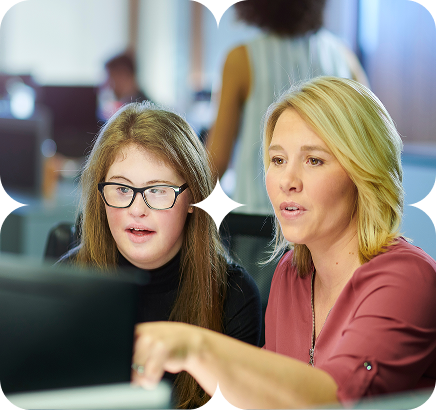 A young woman with glasses and a woman with blonde hair look at a computer screen together in an office, one pointing at the screen.