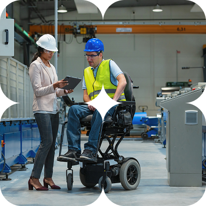 A woman in a white hard hat holds a tablet while talking with a man in a blue hard hat and hi-vis vest who uses a wheelchair, in a warehouse.