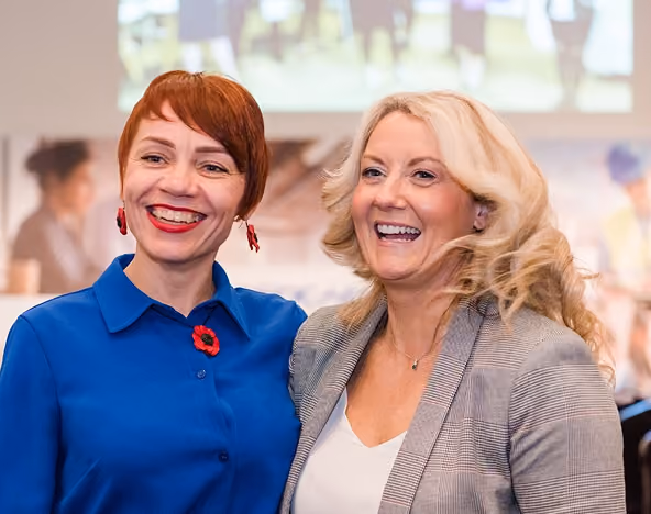 Two smiling women standing close together at an event, one in a blue shirt with a red poppy pin and one in a grey blazer.