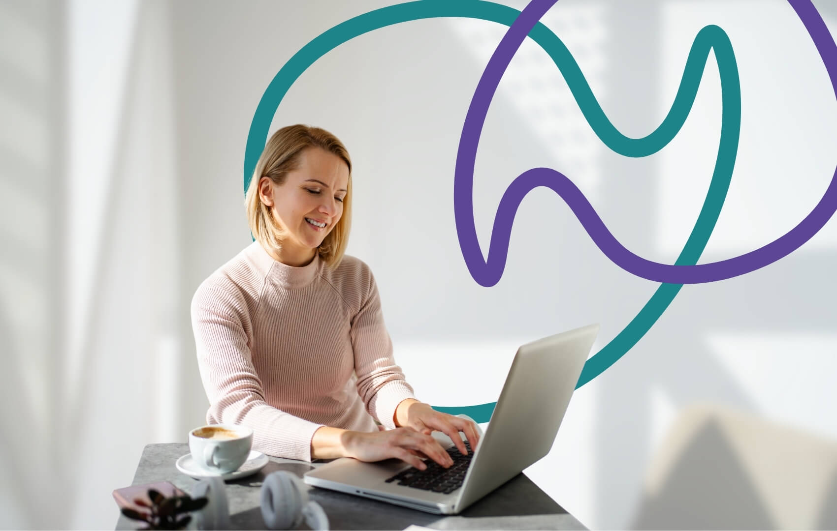 A woman sits at a desk with a laptop and a coffee, smiling as she types.