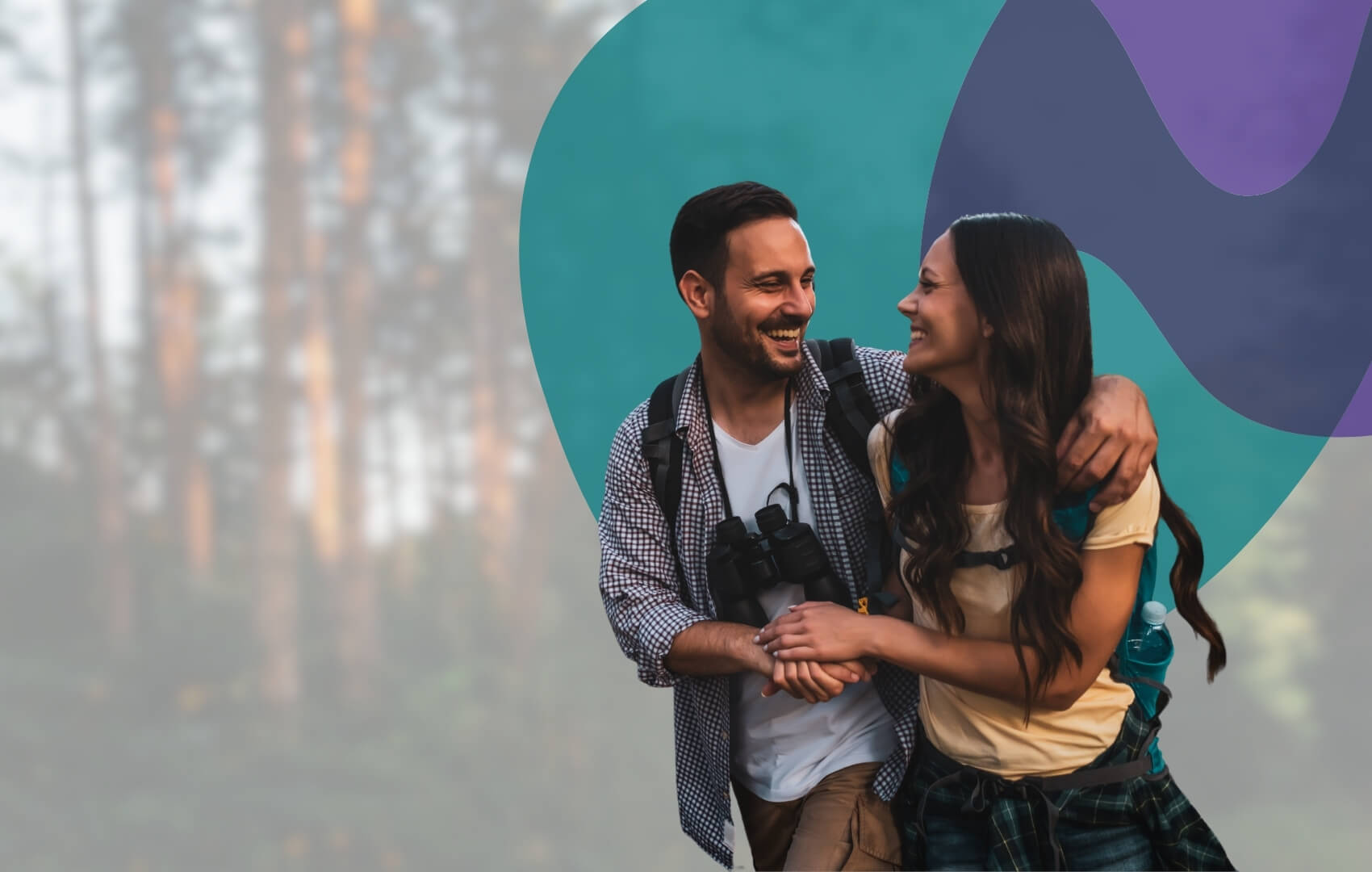 A couple laugh together outdoors, wearing hiking gear and holding each other close. 