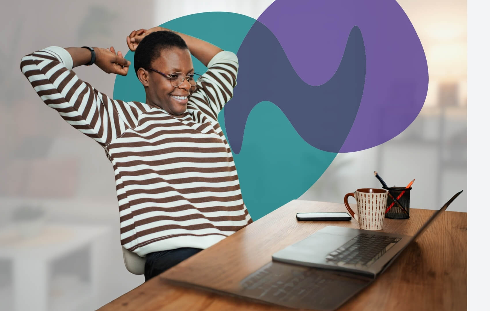 A person sits at a desk with a laptop, stretching their arms back and smiling.