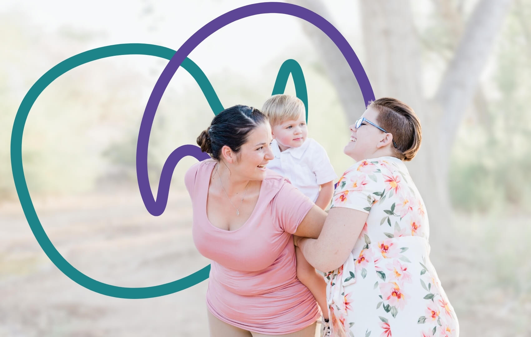 Two mums stand outdoors holding a young toddler between them, smiling at each other and the child.
