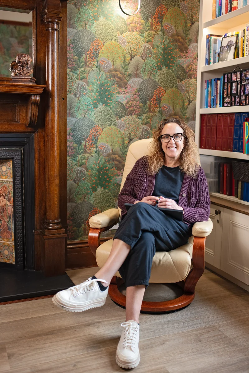 Michelle, a white woman with curly auburn hair and glasses, wearing a purple cardigan, sits in a cream leather chair in a cosy therapy room with a decorative forest wallpaper, wooden fireplace, and bookshelf.