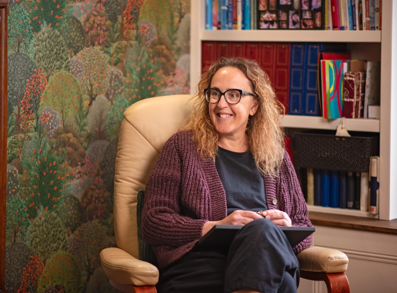 Michelle, a white woman with curly auburn hair and glasses, wearing a purple cardigan, sits smiling in a cream leather chair holding a tablet, with a colourful forest wallpaper and bookshelf visible behind her.