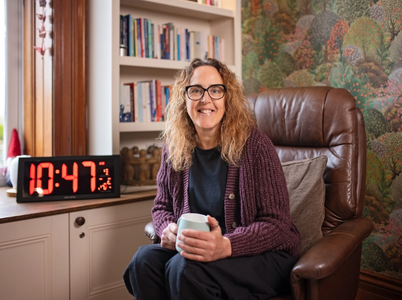 Michelle, a white woman with curly auburn hair and glasses, sits smiling in a brown leather chair holding a mug, with a bookshelf, large digital clock, and decorative wallpaper visible behind her.