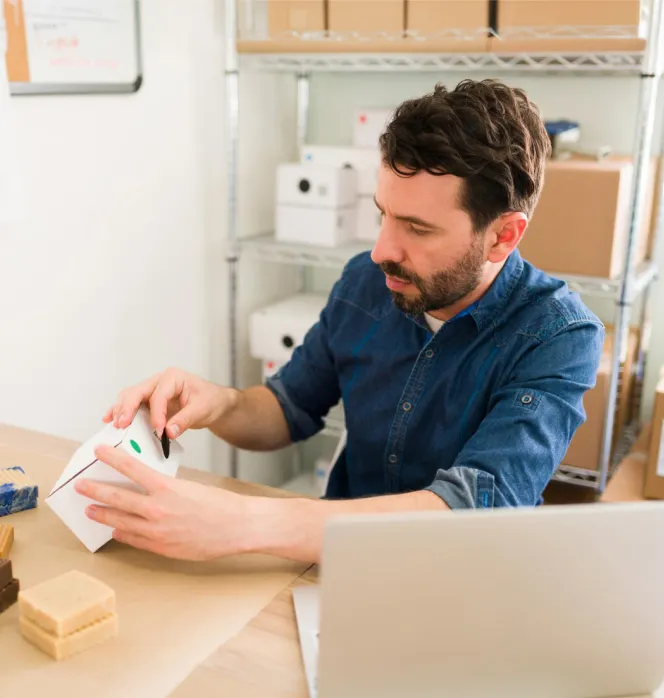 Man in a denim shirt assembling or inspecting a small white box at a cluttered workspace with a laptop and shelving in the background.