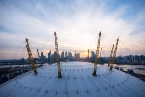 Aerial view of large white dome structure with construction cranes and city skyline at sunset.