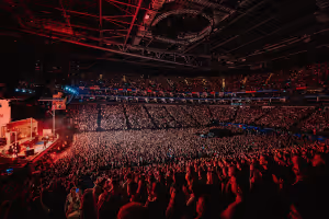 Packed indoor arena crowd watching a concert with red stage lighting and dark ceiling.