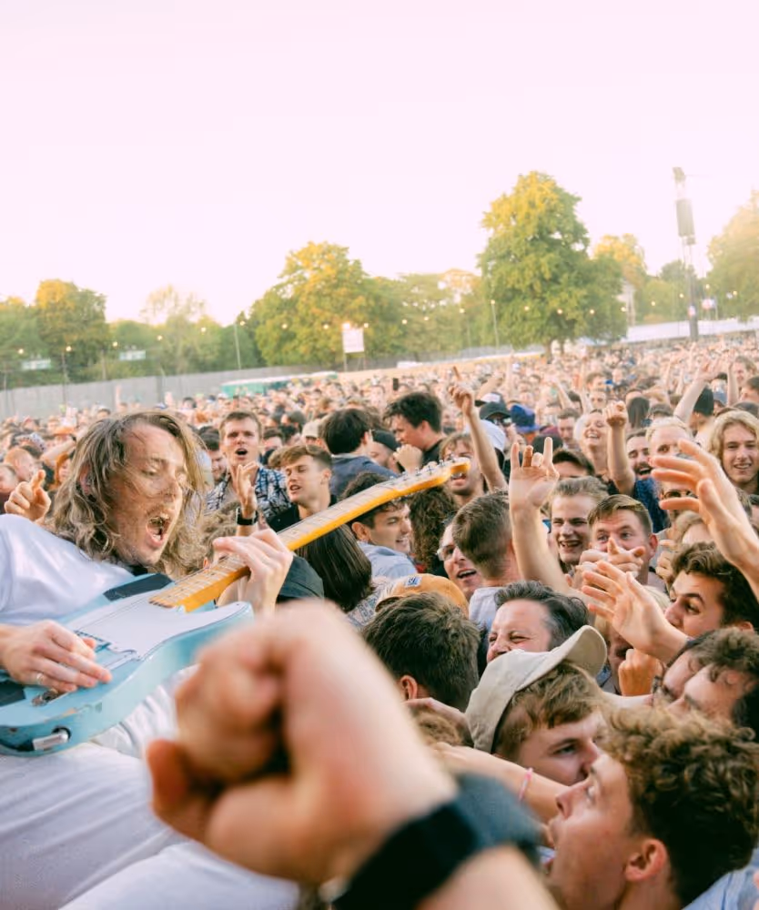 Electric guitarist crowd surfing while energetic audience cheers and raises hands outdoors.