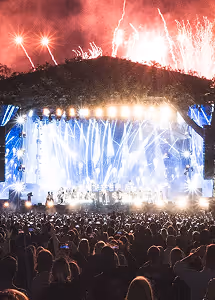 Night concert with bright stage lights, large crowd, and fireworks above a hillside.