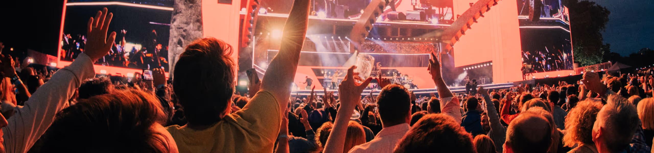 Crowd at outdoor concert cheering with hands raised in front of brightly lit stage with musicians.