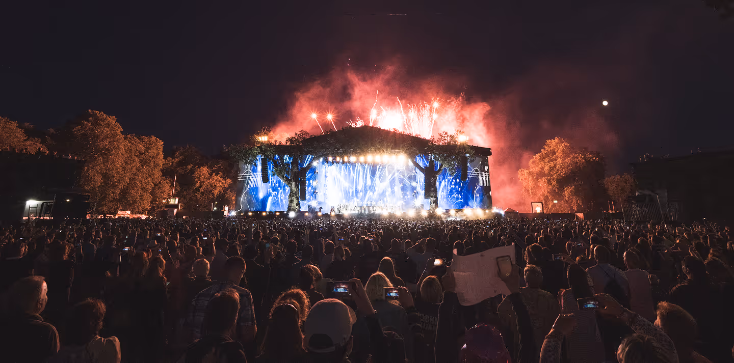Large crowd at night watching a brightly lit outdoor concert stage with fireworks in the sky.