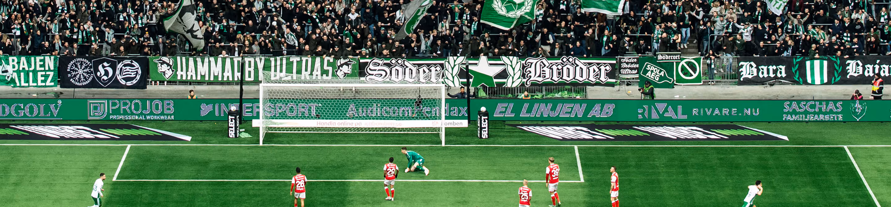 Soccer penalty kick in progress with goalkeeper on ground and crowd waving green and white flags.