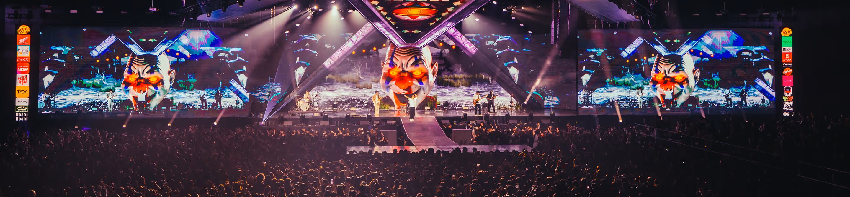 Concert crowd watching band perform on stage with giant illuminated clown faces on screens.