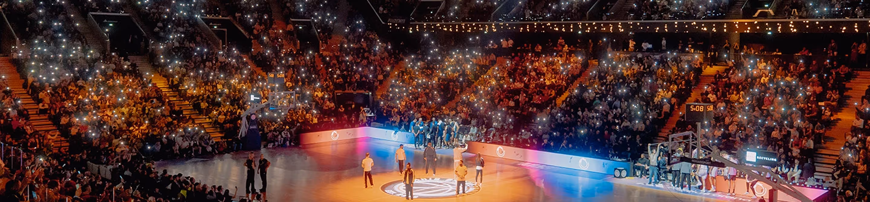 Indoor basketball court with a central team huddle and many spectators holding up illuminated phones.
