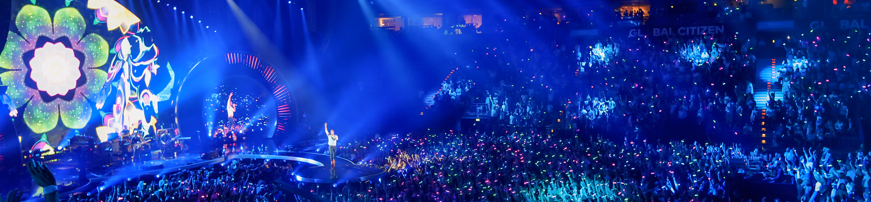 Performers on stage at a concert with large crowd holding colorful glow sticks under blue lights.