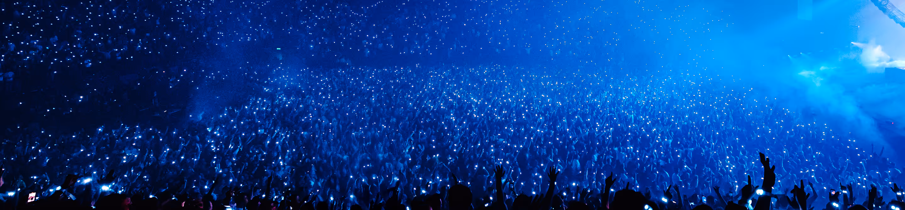 Large crowd at a concert holding up lights in a blue-lit arena with raised hands.