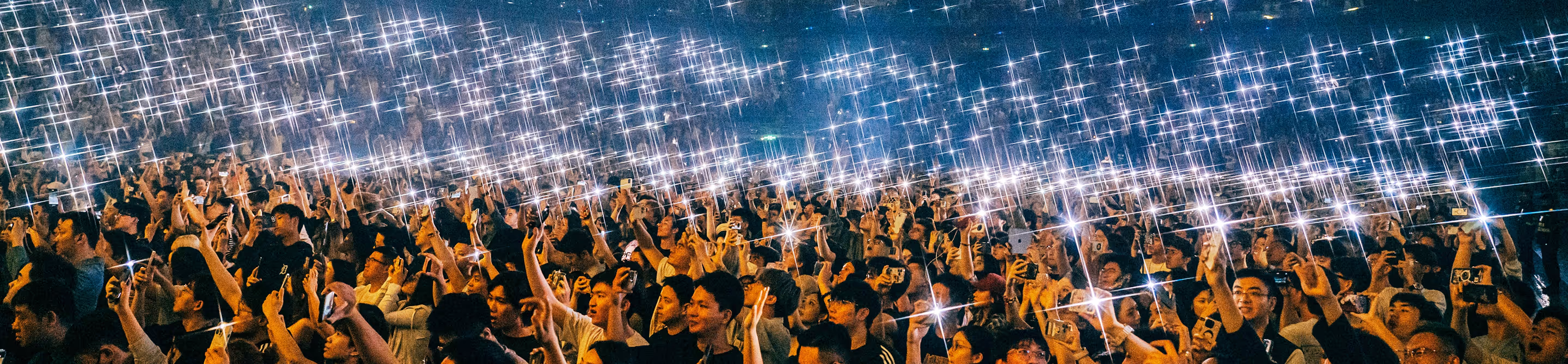 Large crowd holding smartphones with flashlights on, creating a sparkling light effect in the night.