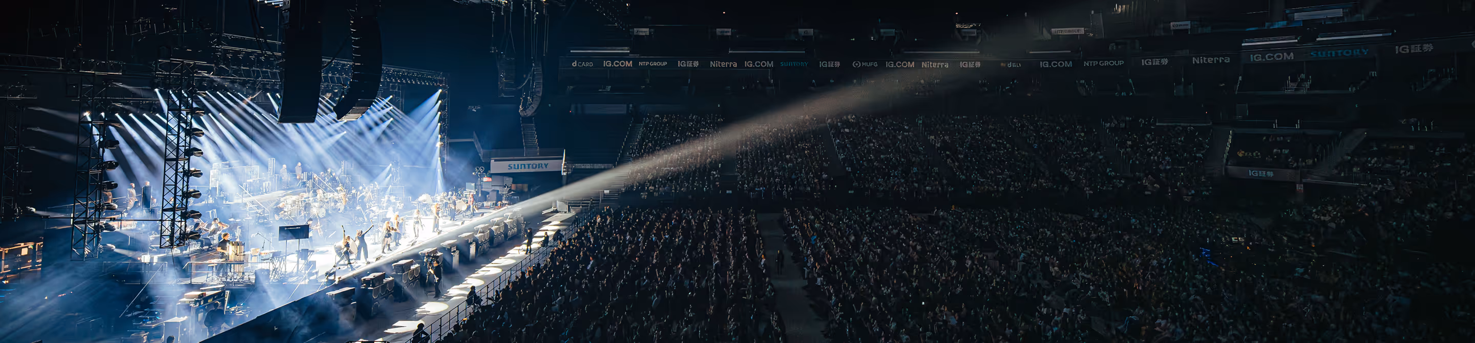 Wide shot of a concert stage with performers amid bright lights and a large dark audience.