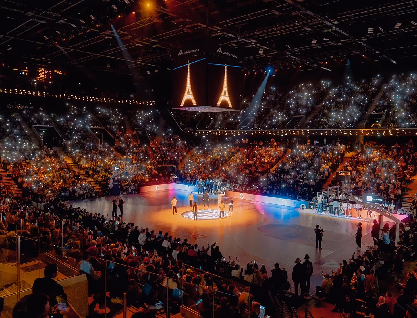 Indoor basketball arena filled with spectators holding up lights and a lit Eiffel Tower display above the court.
