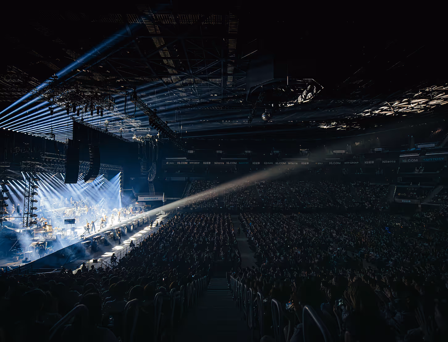 Concert stage with bright spotlights shining on performers before a large seated audience in a dark arena.