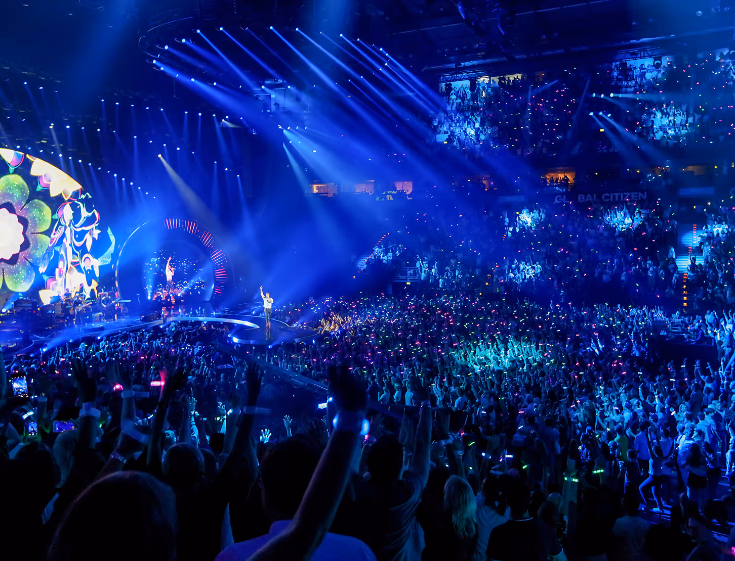 Crowd cheering at a concert with blue stage lights and musicians performing on a large stage.