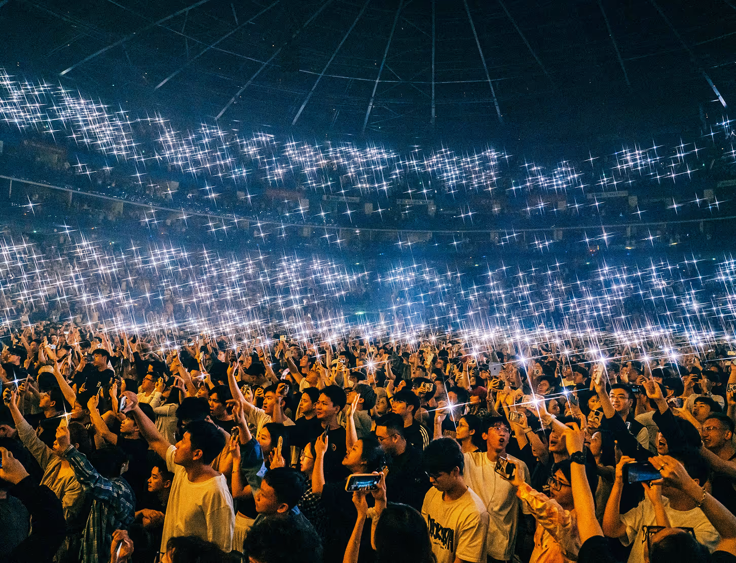 Crowd at a concert holding up lit phones creating a sparkling light effect in a large indoor arena.