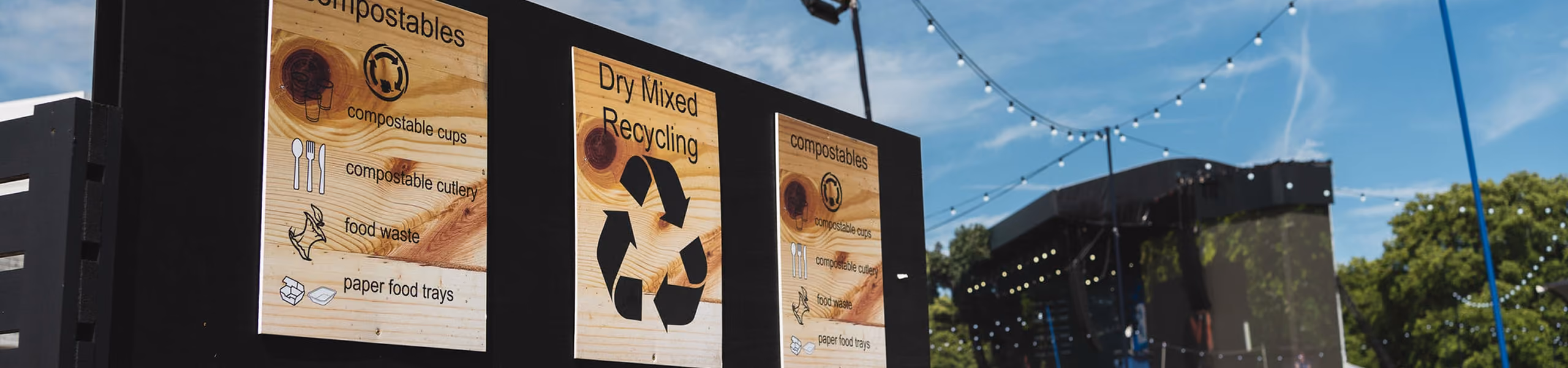 Wooden recycling signs for compostables and dry mixed recycling at an outdoor event with string lights.
