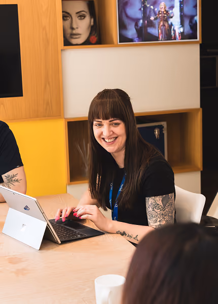 Smiling woman with tattoos typing on a laptop at a meeting table in a modern office.