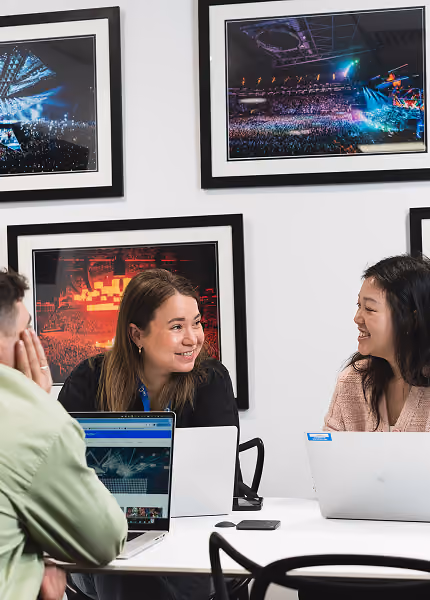 Three people smiling and chatting around a table with laptops in a room with framed concert photos.