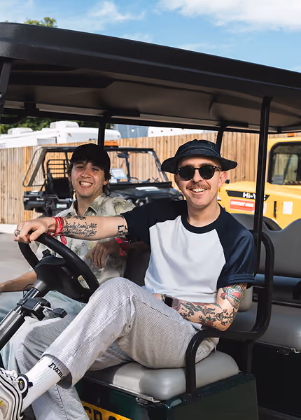 Two men sitting in a golf cart, one wearing sunglasses and a bucket hat with tattoos on his arms.