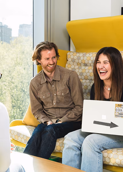 Two people laughing while sitting on a yellow and patterned sofa with a laptop on the woman's lap.