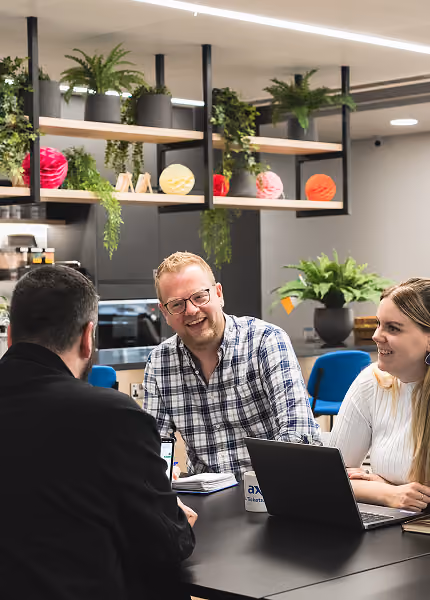 Three people smiling and talking around a table with laptops in a modern office space with plants.