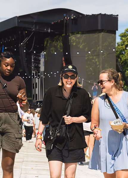 Three young women smiling and walking outdoors near a festival stage on a sunny day.
