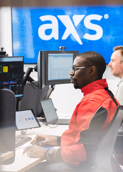 Two men working at desks with computers in front of a blue screen displaying the axs logo.
