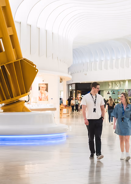 Two people walking inside a bright, modern mall with white curved ceiling and large yellow sculpture.